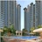 Tall modern residential buildings with numerous windows rise against a clear sky. In the foreground, a swimming pool is surrounded by lounge chairs and umbrellas. A domed pavilion and lush green trees add to the serene setting. A person stands near the pool area.