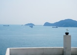 A seascape featuring a few cargo ships spread across the calm blue ocean waters. Islands are visible on the horizon under a hazy sky. A small white structure with a black and yellow device, possibly a weather vane or antenna, is positioned in the foreground.