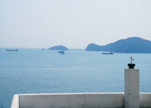A seascape featuring a few cargo ships spread across the calm blue ocean waters. Islands are visible on the horizon under a hazy sky. A small white structure with a black and yellow device, possibly a weather vane or antenna, is positioned in the foreground.