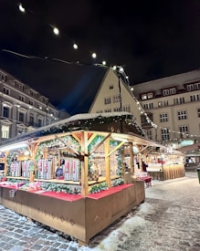 A festive market stall adorned with holiday lights and decorations, situated in a snow-covered area among historic buildings. The stall's structure is decorated with garlands and red fabric, and it displays various items like beverages or food products. The scene is illuminated by strings of lights, and a cozy, inviting atmosphere is evident.