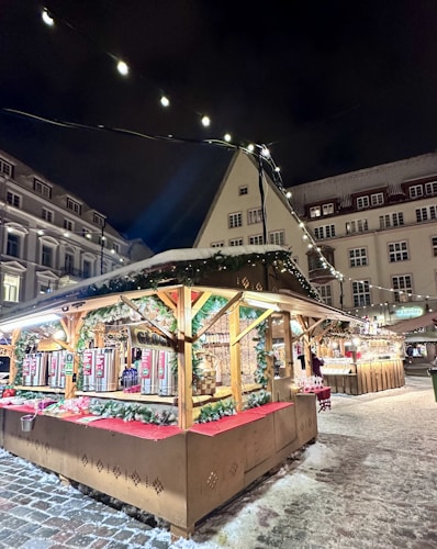 A festive market stall adorned with holiday lights and decorations, situated in a snow-covered area among historic buildings. The stall's structure is decorated with garlands and red fabric, and it displays various items like beverages or food products. The scene is illuminated by strings of lights, and a cozy, inviting atmosphere is evident.