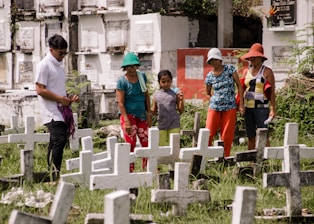A group of five people stand among multiple white crosses in a grassy cemetery. They appear to be paying their respects at the gravesites. The background features above-ground burial niches in white with inscriptions, and the people are casually dressed with hats to shield from the sun.