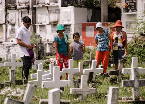 A group of five people stand among multiple white crosses in a grassy cemetery. They appear to be paying their respects at the gravesites. The background features above-ground burial niches in white with inscriptions, and the people are casually dressed with hats to shield from the sun.