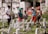 A group of five people stand among multiple white crosses in a grassy cemetery. They appear to be paying their respects at the gravesites. The background features above-ground burial niches in white with inscriptions, and the people are casually dressed with hats to shield from the sun.