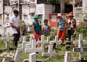 A group of five people stand among multiple white crosses in a grassy cemetery. They appear to be paying their respects at the gravesites. The background features above-ground burial niches in white with inscriptions, and the people are casually dressed with hats to shield from the sun.
