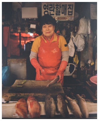 An elderly person wearing a yellow shirt and a red apron and gloves stands behind a counter, holding a fish. The counter displays various fish, and the background includes hanging items, plastic bags, a sign with Korean text, and a somewhat dimly lit setting.
