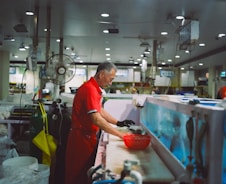 A man in a red uniform is working at a seafood market stall, cleaning fish at a counter. The space is filled with various equipment, including oxygen tanks and overhead lights. A fan is mounted above his workstation, and there are fish tanks visible in the background.