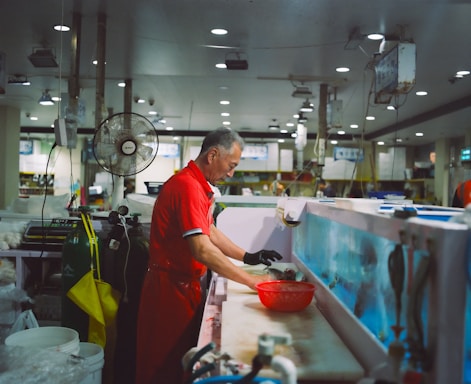 A man in a red uniform is working at a seafood market stall, cleaning fish at a counter. The space is filled with various equipment, including oxygen tanks and overhead lights. A fan is mounted above his workstation, and there are fish tanks visible in the background.