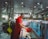 A man in a red uniform is working at a seafood market stall, cleaning fish at a counter. The space is filled with various equipment, including oxygen tanks and overhead lights. A fan is mounted above his workstation, and there are fish tanks visible in the background.