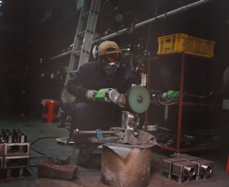 A person working in a workshop, wearing protective gear, operates a power tool on a metal piece. The environment includes various metal parts, a ladder, and a fan in the background. A yellow plastic basket and red shelving are visible.