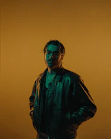 A model wearing a dark emerald green leather jacket with gold accents, standing against a textured stone wall.