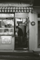 A small shop with a striped awning and various items displayed in the window. A person is seen inside, standing and looking down at something. The shop has signage in both Korean and English indicating its hours of operation. There is an ADT security sign on the wall.
