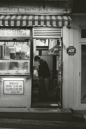 A small shop with a striped awning and various items displayed in the window. A person is seen inside, standing and looking down at something. The shop has signage in both Korean and English indicating its hours of operation. There is an ADT security sign on the wall.