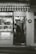 A small shop with a striped awning and various items displayed in the window. A person is seen inside, standing and looking down at something. The shop has signage in both Korean and English indicating its hours of operation. There is an ADT security sign on the wall.
