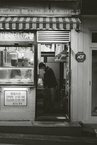 A small shop with a striped awning and various items displayed in the window. A person is seen inside, standing and looking down at something. The shop has signage in both Korean and English indicating its hours of operation. There is an ADT security sign on the wall.