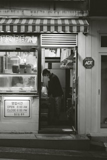 A small shop with a striped awning and various items displayed in the window. A person is seen inside, standing and looking down at something. The shop has signage in both Korean and English indicating its hours of operation. There is an ADT security sign on the wall.