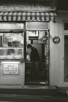 A small shop with a striped awning and various items displayed in the window. A person is seen inside, standing and looking down at something. The shop has signage in both Korean and English indicating its hours of operation. There is an ADT security sign on the wall.