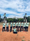 A group of individuals, mostly young males, standing on a reddish-brown dirt field. They are wearing light green shirts and dark pants. Two individuals sit on the crossbar of a goalpost, while others stand in front, posing with hands clasped. A bright sky with scattered clouds is visible above, and there are trees in the background.