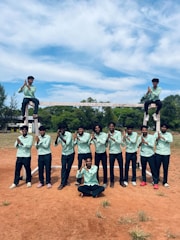 A group of individuals, mostly young males, standing on a reddish-brown dirt field. They are wearing light green shirts and dark pants. Two individuals sit on the crossbar of a goalpost, while others stand in front, posing with hands clasped. A bright sky with scattered clouds is visible above, and there are trees in the background.