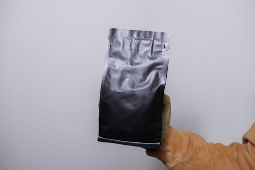 A hand is holding a black, matte-finish, resealable pouch against a plain white background. The pouch is smooth and simple, indicating it may be used for packaging food or other products.
