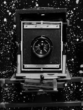 A vintage large format camera with a prominent lens is centered against a speckled, star-like black and white background. The camera features a sturdy metal frame and a bellows design, typical of early photographic equipment.