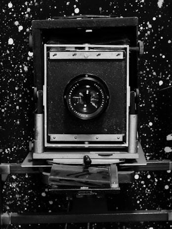 A vintage large format camera with a prominent lens is centered against a speckled, star-like black and white background. The camera features a sturdy metal frame and a bellows design, typical of early photographic equipment.
