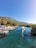A serene riverside scene with several small boats moored along the bank. A boat with a canopy is moving through the clear, blue-green water, creating a gentle wake. The backdrop features lush greenery and a majestic mountain under a clear blue sky.