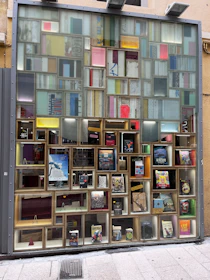 A colorful display window with an assortment of books and framed art pieces arranged in a grid-like pattern. The window features various book covers, artwork, and decorative items. Some frames are empty, adding to the abstract geometric design.
