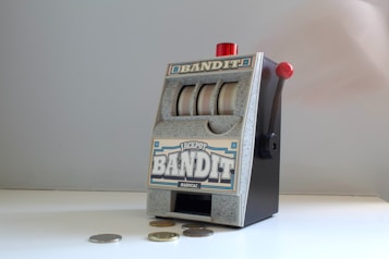 A small, vintage-style slot machine with the word 'Bandit' prominently displayed. It features a lever on the side with a red knob and three reels with a generic design. Several coins are scattered on the white surface in front of it.