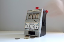 A small, vintage-style slot machine with the word 'Bandit' prominently displayed. It features a lever on the side with a red knob and three reels with a generic design. Several coins are scattered on the white surface in front of it.