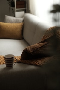 A cozy, neutral-colored sofa is accentuated by textured brown pillows and a soft blanket. A cup of coffee in a striped mug is placed on the sofa, adding a touch of warmth and relaxation. The soft light filtering through a window creates a tranquil and inviting atmosphere.