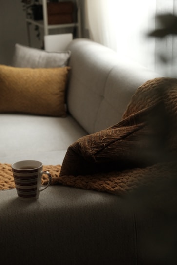A cozy, neutral-colored sofa is accentuated by textured brown pillows and a soft blanket. A cup of coffee in a striped mug is placed on the sofa, adding a touch of warmth and relaxation. The soft light filtering through a window creates a tranquil and inviting atmosphere.
