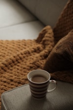 A cozy setting featuring a striped mug filled with a warm beverage placed on an armrest. A textured brown blanket covers a sofa, creating a warm and inviting atmosphere.