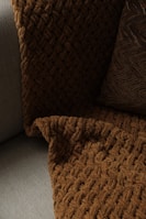 A close-up view of a textured, woven blanket and a pillow placed on a couch. The blanket has a rich, brown color with a chunky knit design, while the pillow features a similar dark tone with a zigzag pattern. The fabric contrasts softly against the lighter gray color of the couch upholstery.