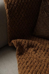 A close-up view of a textured, woven blanket and a pillow placed on a couch. The blanket has a rich, brown color with a chunky knit design, while the pillow features a similar dark tone with a zigzag pattern. The fabric contrasts softly against the lighter gray color of the couch upholstery.