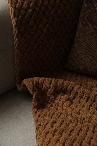 A close-up view of a textured, woven blanket and a pillow placed on a couch. The blanket has a rich, brown color with a chunky knit design, while the pillow features a similar dark tone with a zigzag pattern. The fabric contrasts softly against the lighter gray color of the couch upholstery.