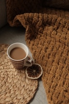 A cozy setting with a textured brown knit blanket draped over a surface. A woven mat lies beside a striped mug filled with coffee or hot chocolate. A dried citrus slice adds a decorative touch.