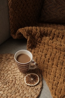 A cozy setting featuring a striped mug filled with coffee or tea, placed on a woven round placemat alongside a slice of dried citrus. An earthy-toned knitted blanket is draped over a couch.