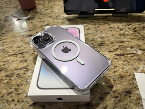 A smartphone with a clear protective case is placed on top of its box on a speckled granite countertop. The case features a circular design around a logo. In the background, a larger device screen is visible, set at an angle.