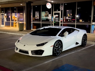 A sleek, white sports car is parked in front of a retail establishment with large glass windows. The area is illuminated by artificial street and store lighting, highlighting the car’s glossy finish and distinctive wheel design. Various store signs and posters are visible in the background, suggesting a commercial area.