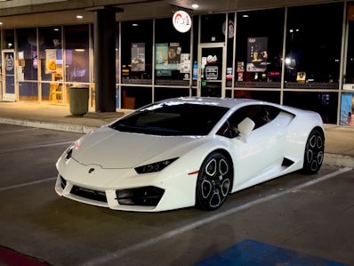 A sleek, white sports car is parked in front of a retail establishment with large glass windows. The area is illuminated by artificial street and store lighting, highlighting the car’s glossy finish and distinctive wheel design. Various store signs and posters are visible in the background, suggesting a commercial area.