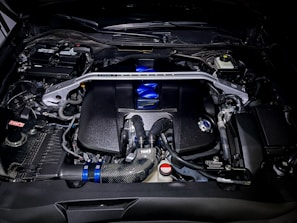 A detailed view of a high-performance car engine, featuring various components such as pipes, valves, and a distinct metallic support bar across the top. The engine has a polished look with elements in black, blue, and silver. Various branding and labels are visible on different parts.