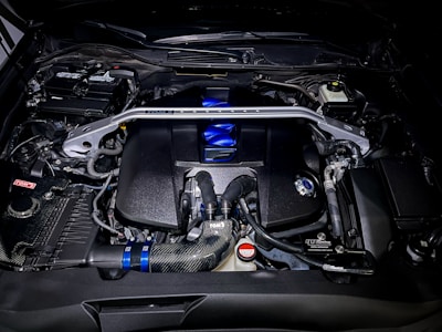A detailed view of a high-performance car engine, featuring various components such as pipes, valves, and a distinct metallic support bar across the top. The engine has a polished look with elements in black, blue, and silver. Various branding and labels are visible on different parts.