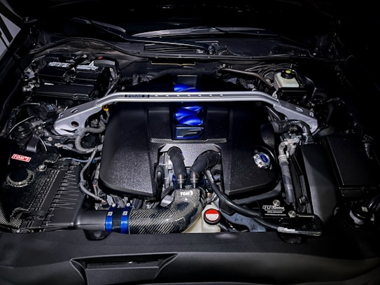 A detailed view of a high-performance car engine, featuring various components such as pipes, valves, and a distinct metallic support bar across the top. The engine has a polished look with elements in black, blue, and silver. Various branding and labels are visible on different parts.