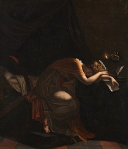 A woman dressed in a flowing, dark gown leans over a table in a dimly lit room. Her face is partially obscured by her arm resting on the table, and she holds a scroll in her other hand. An oil lamp burns nearby, casting a warm glow over her, adding a sense of mystery and introspection.