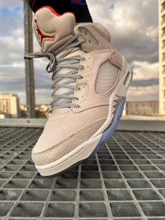 Close-up of a light-colored sneaker with detailed stitching and a mesh side panel. The shoe features a red logo on the tongue and gray laces with a checkered pattern. The background includes a metallic grid floor and a blurred cityscape with buildings visible.