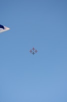 A formation of red airplanes flying in a tight group against a clear blue sky. The tip of an aircraft wing can be seen on the left side of the image.