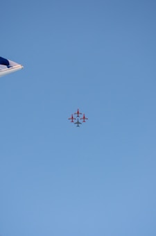 A formation of red airplanes flying in a tight group against a clear blue sky. The tip of an aircraft wing can be seen on the left side of the image.