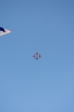 A formation of red airplanes flying in a tight group against a clear blue sky. The tip of an aircraft wing can be seen on the left side of the image.