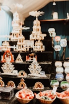 The image depicts a collection of intricate wooden Christmas decorations arranged on black shelves. Among the decorations are large, multi-tiered candle pyramids adorned with small figures and candles. Various other holiday ornaments, such as wooden trees and small nativity scenes, are displayed alongside colorful ceramic candle holders with festive designs. Baskets filled with red and green items are positioned at the bottom of the display.